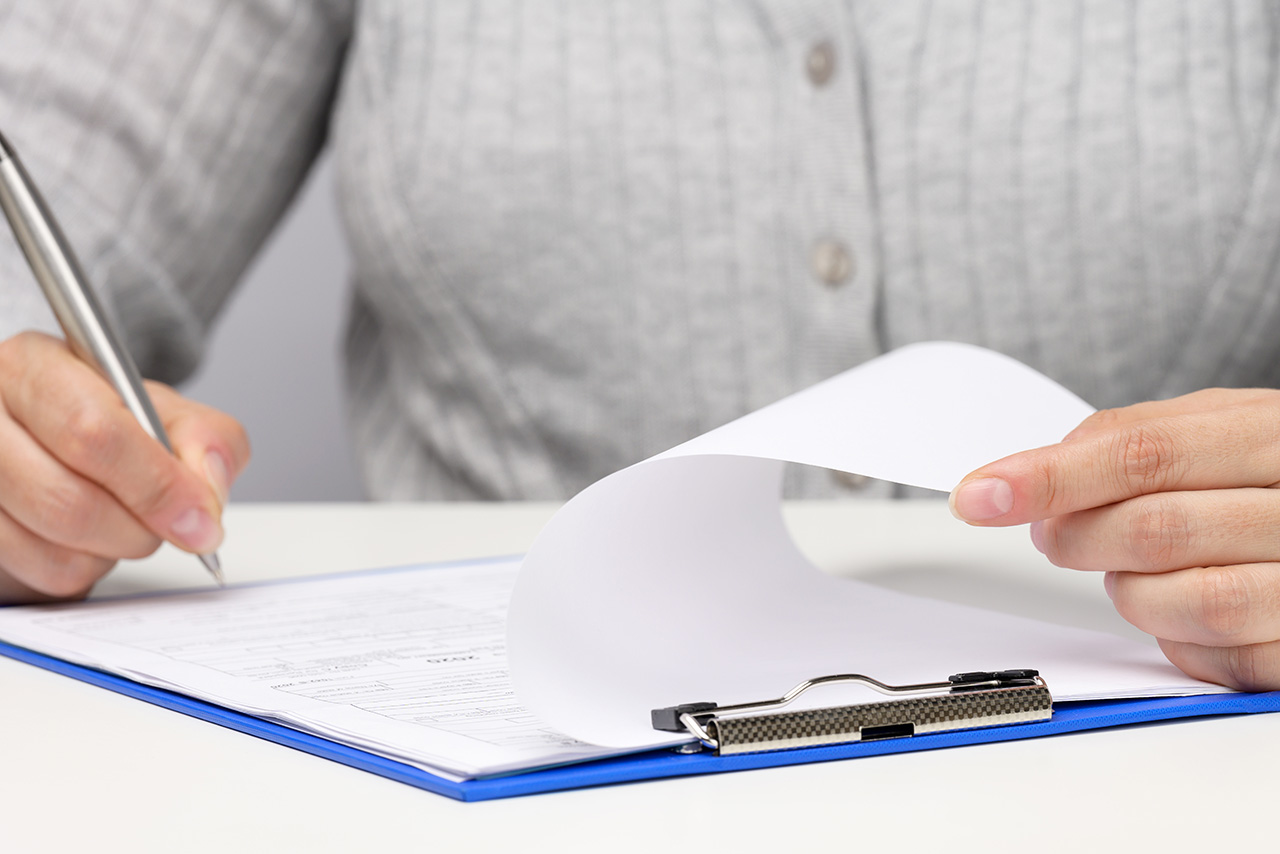 woman-sits-in-gray-clothes woman of Caucasian appearance sits in gray clothes at a white table and signs documents with a metal pen.